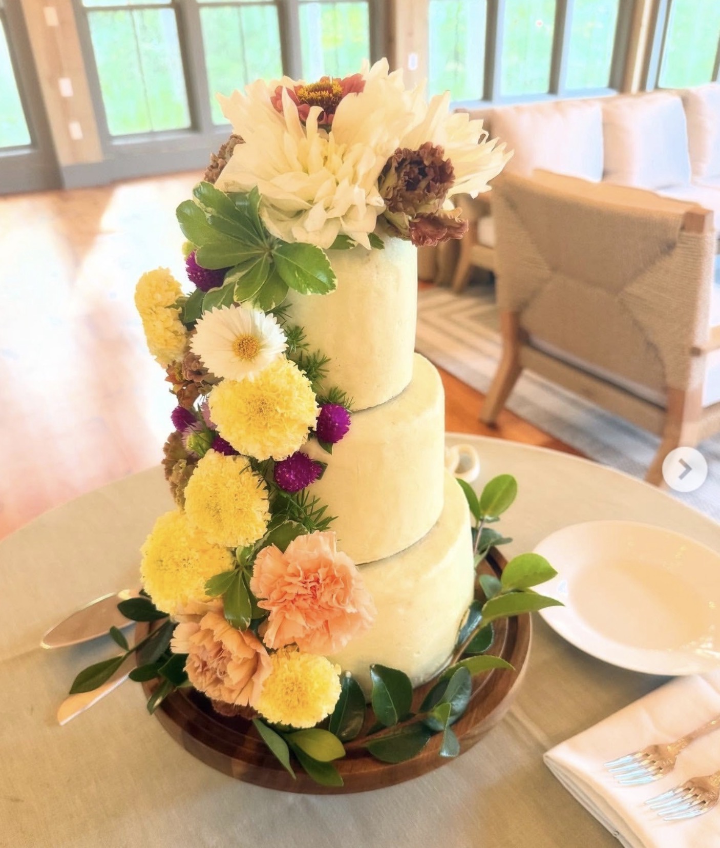 Three-tiered cake decorated with white, yellow, pink, and purple flowers, surrounded by green leaves, on a table in a room.