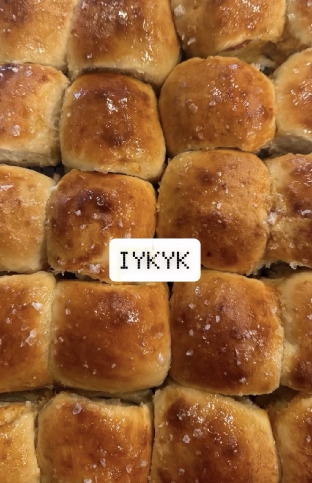 Close-up of golden-brown baked bread rolls with salt crystals, arranged in a grid pattern.