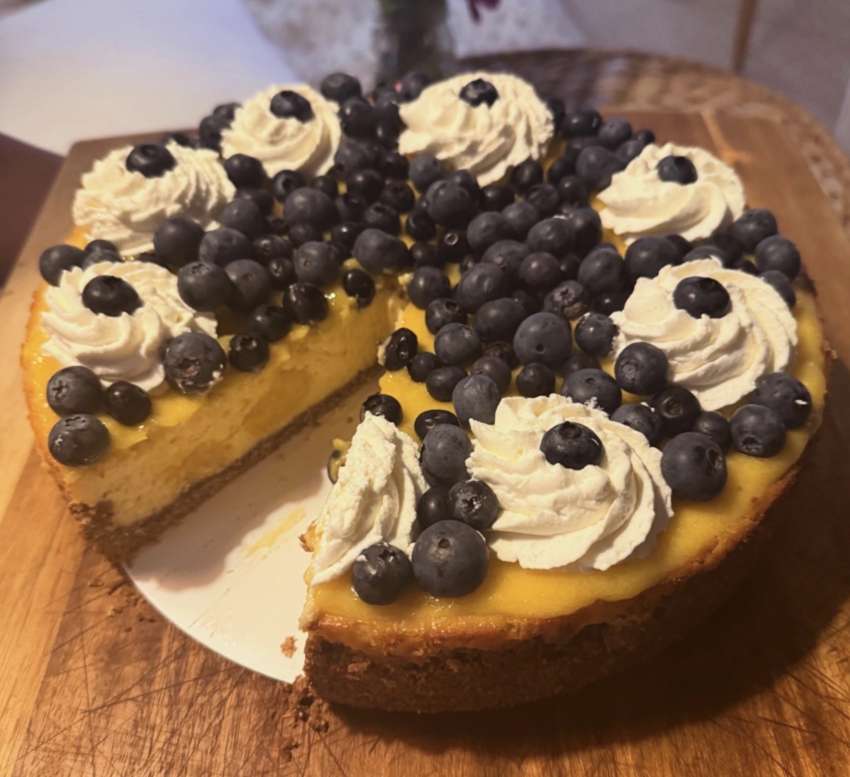 A blueberry-topped cheesecake with whipped cream decorations on a wooden surface, one slice removed.