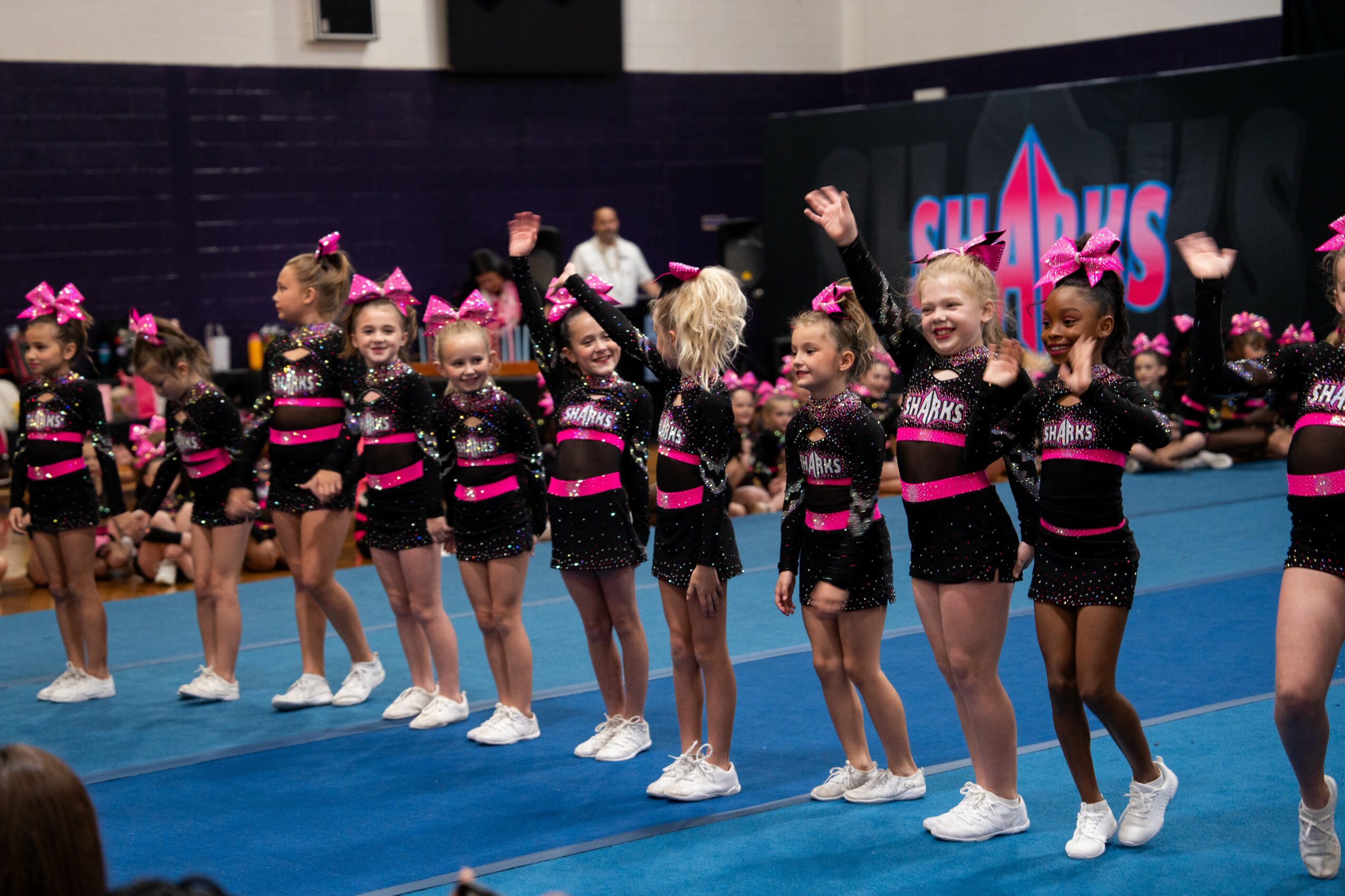 Group of young cheerleaders in black and pink uniforms standing on a blue mat, raising their arms, with a stage and audience in background.