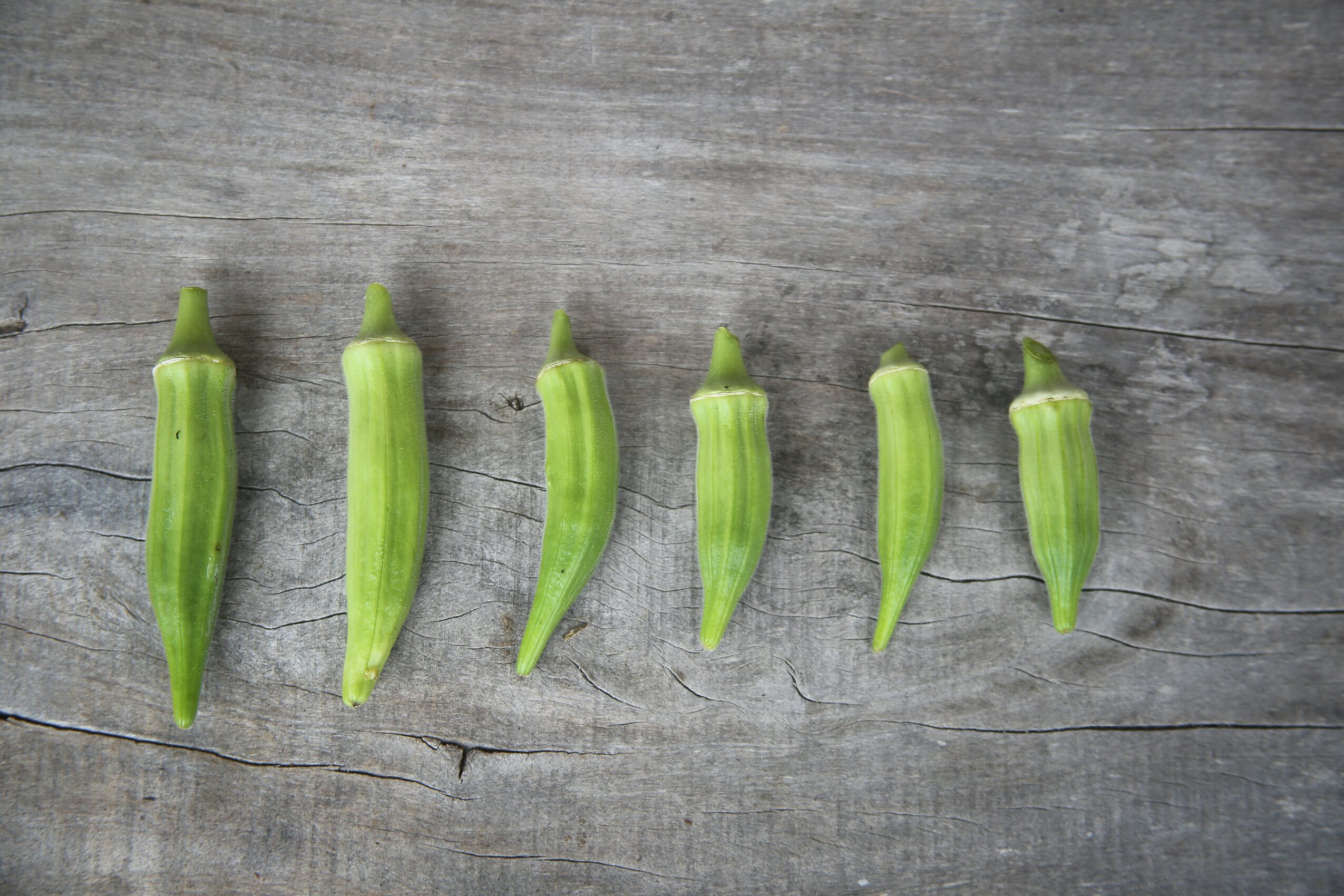 Six green okra pods lined up on a weathered wooden surface, varying slightly in size and shape.