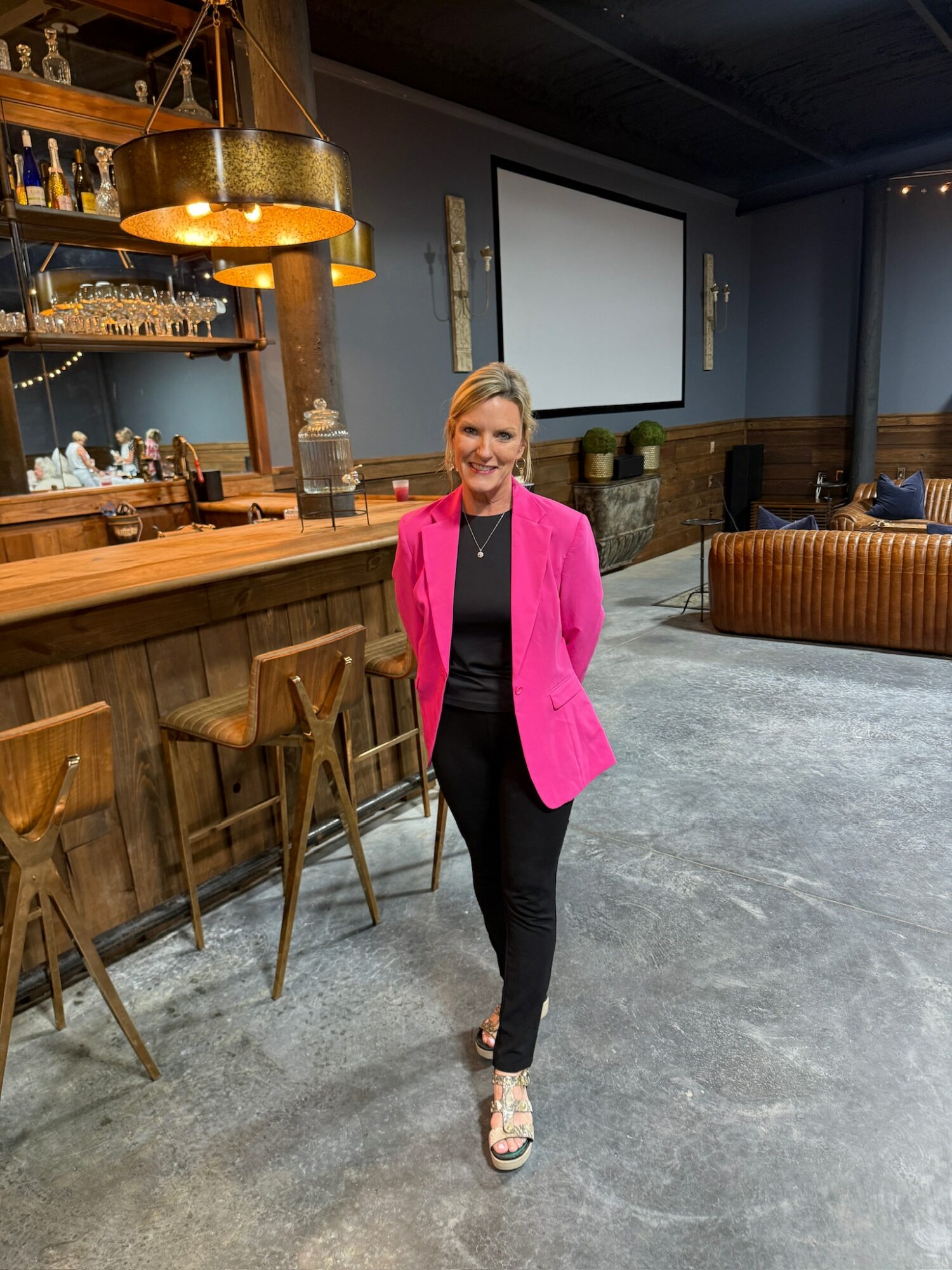 Woman in pink blazer and black pants standing in a bar or lounge area with a wooden bar counter and seating, smiling.