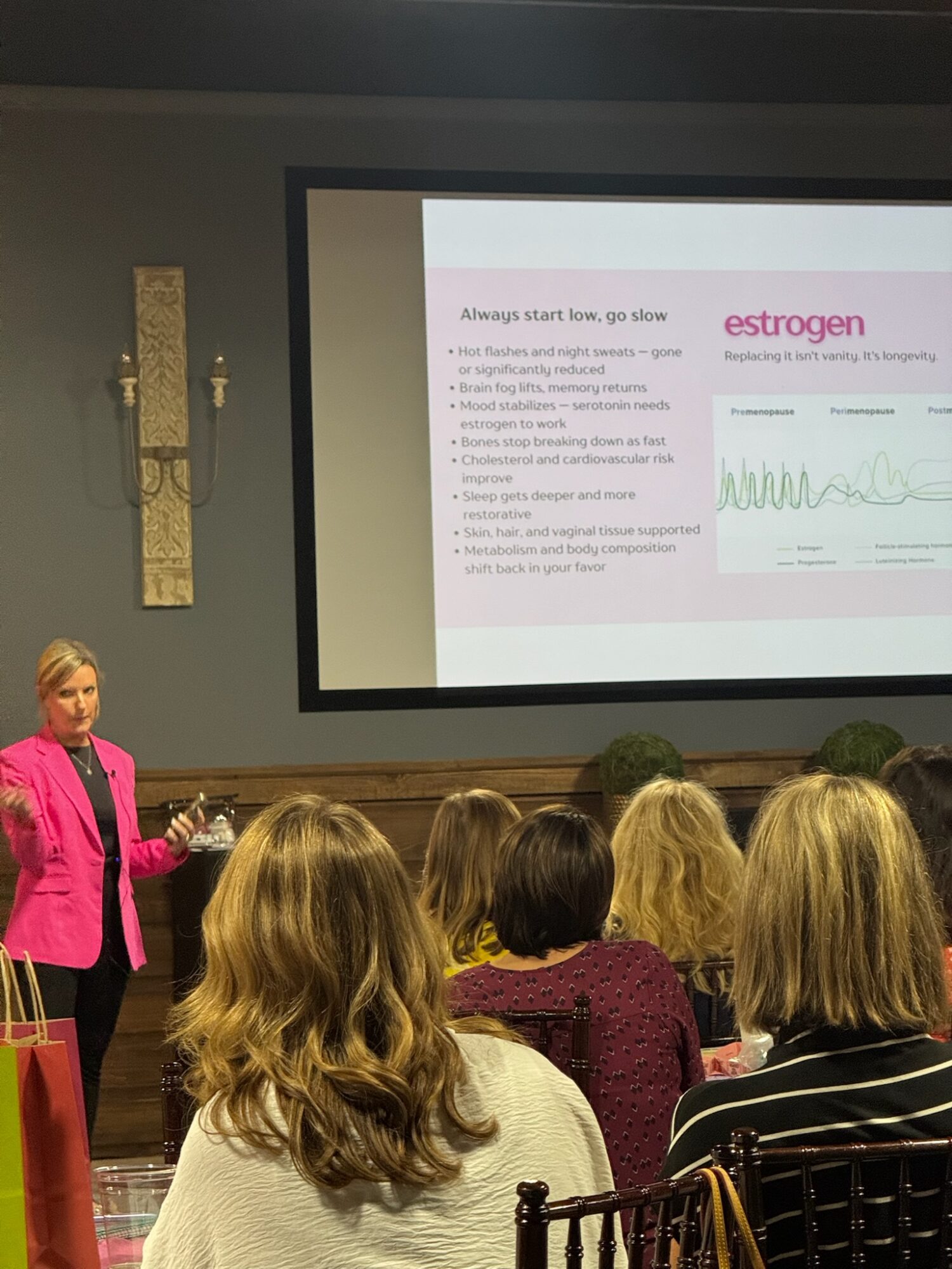A woman in a pink blazer presents to a group of seated people in a conference room with a large screen displaying a presentation about estrogen.