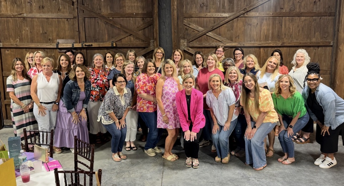 Group of women and men standing together in a large indoor space with wooden walls, smiling and posing for a photo.