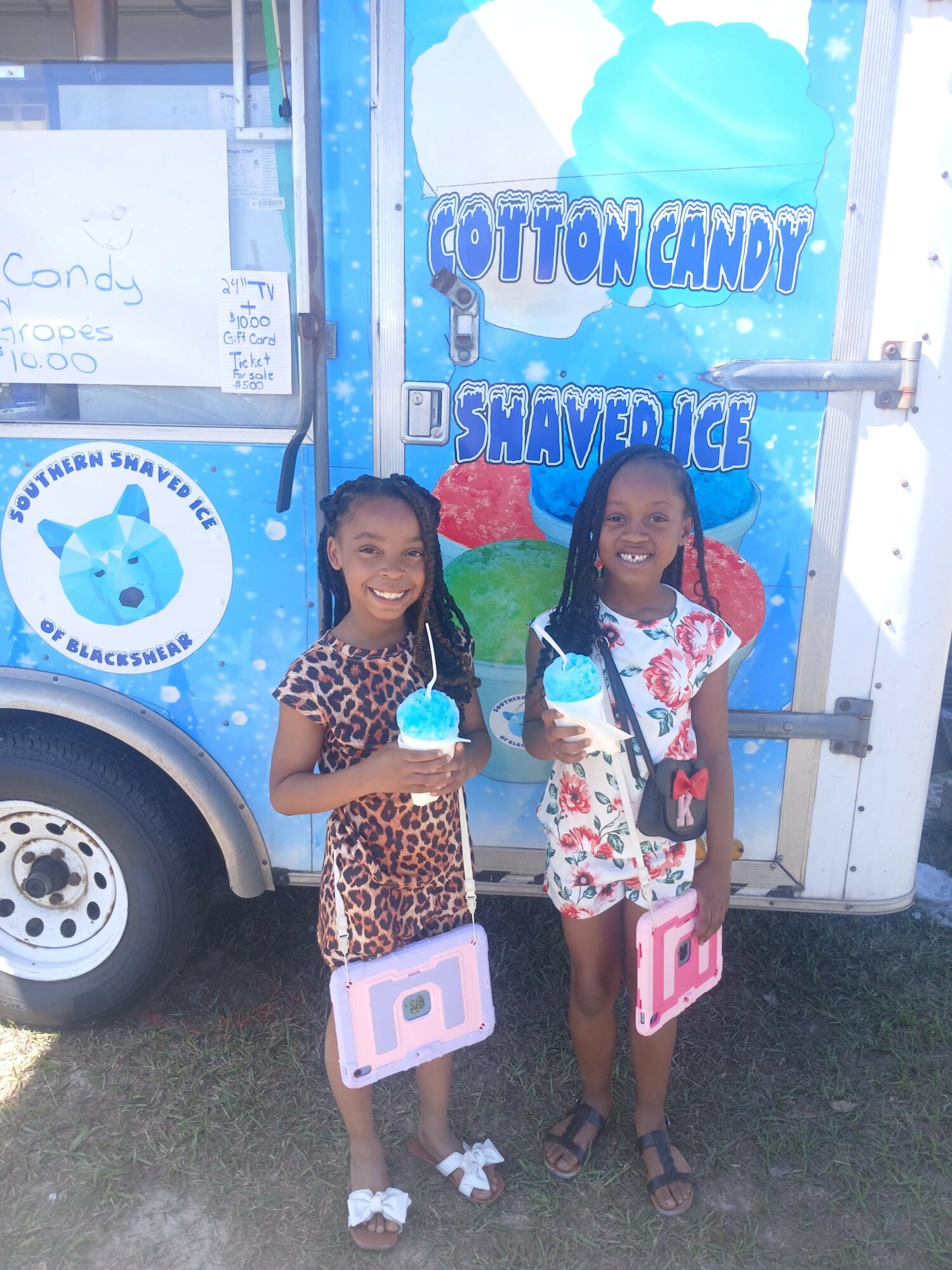 Two young girls stand in front of a cotton candy and shaved ice stand, holding blue treats and pink bags, smiling.