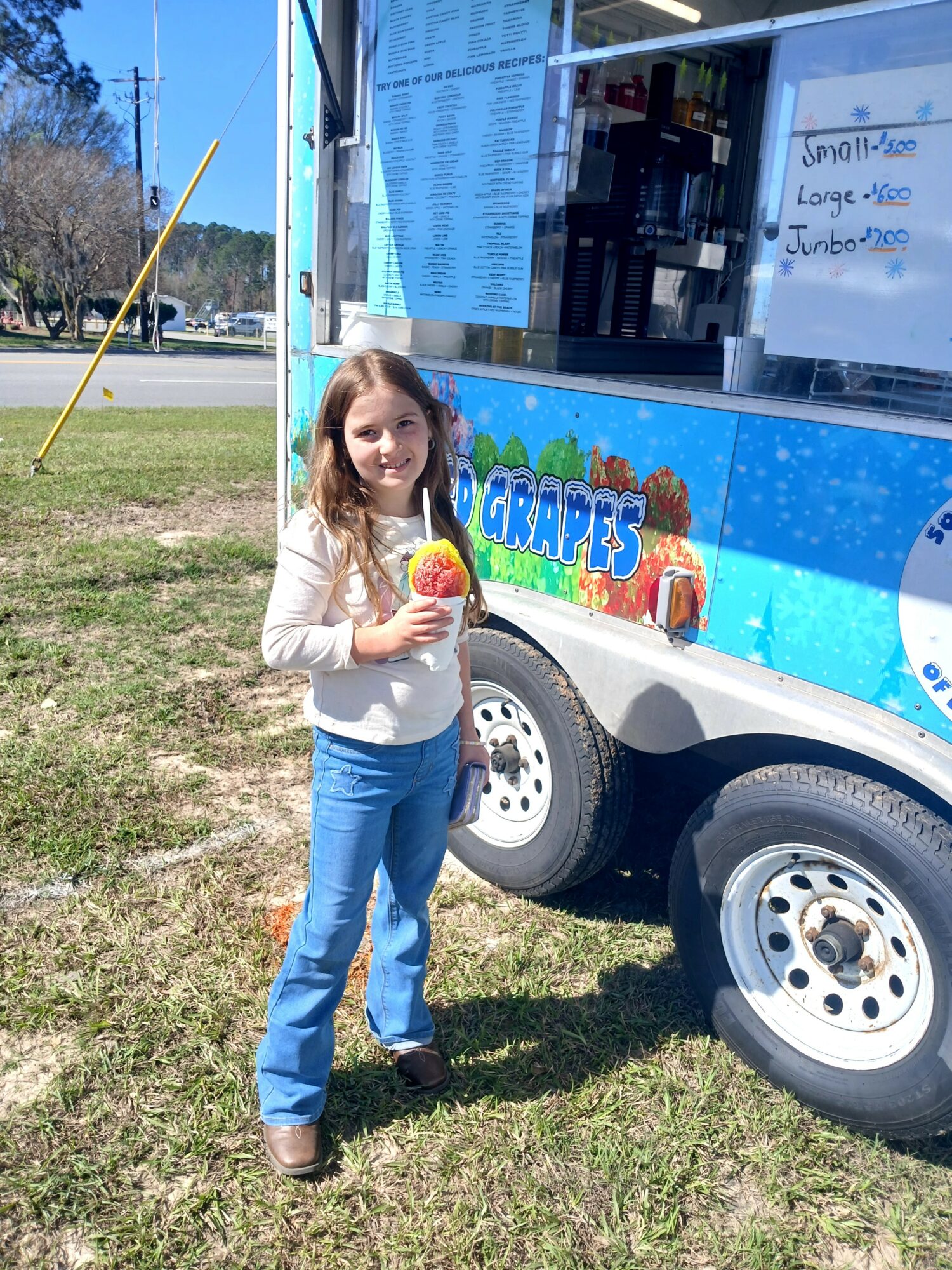 Girl holding a yellow drink in front of a blue food truck with a menu, standing on grass.
