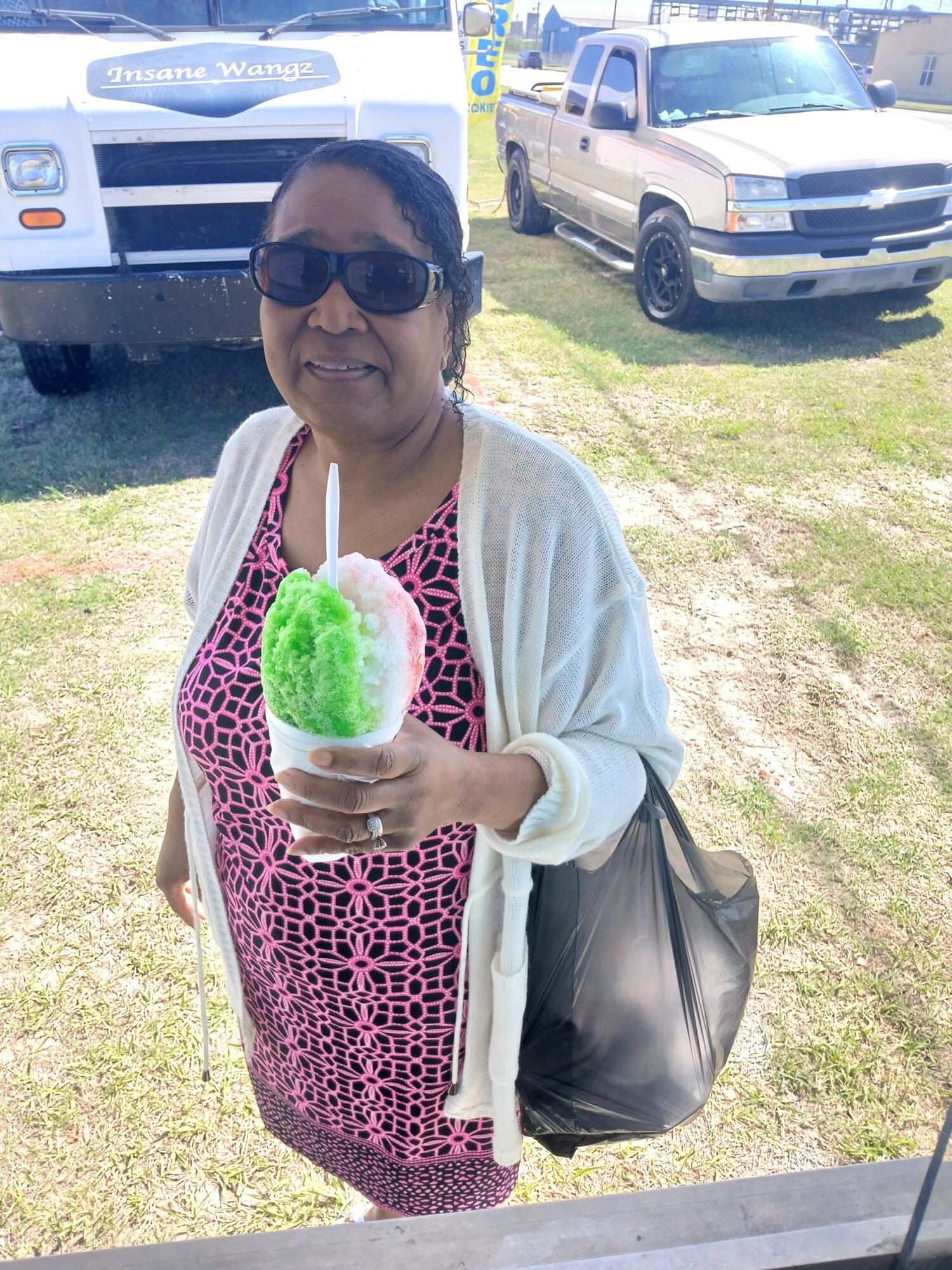 Woman wearing sunglasses holding a green and white cotton candy outdoors, with vehicles in the background.