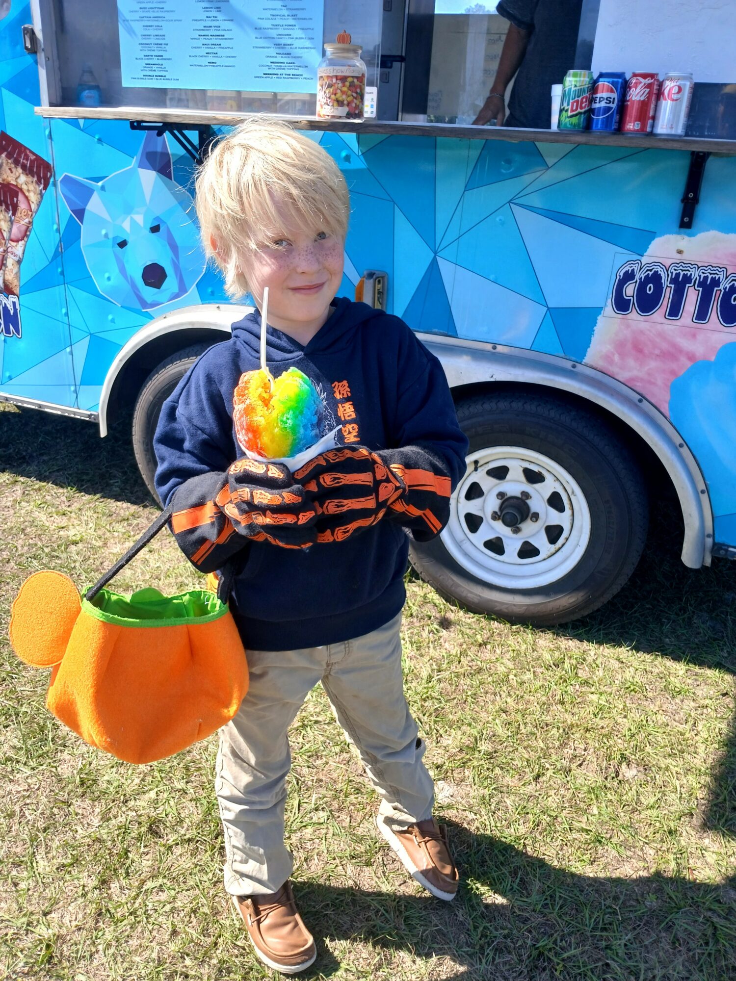 Child holding a rainbow-colored cotton candy in front of a blue ice cream truck, outdoors on grass.