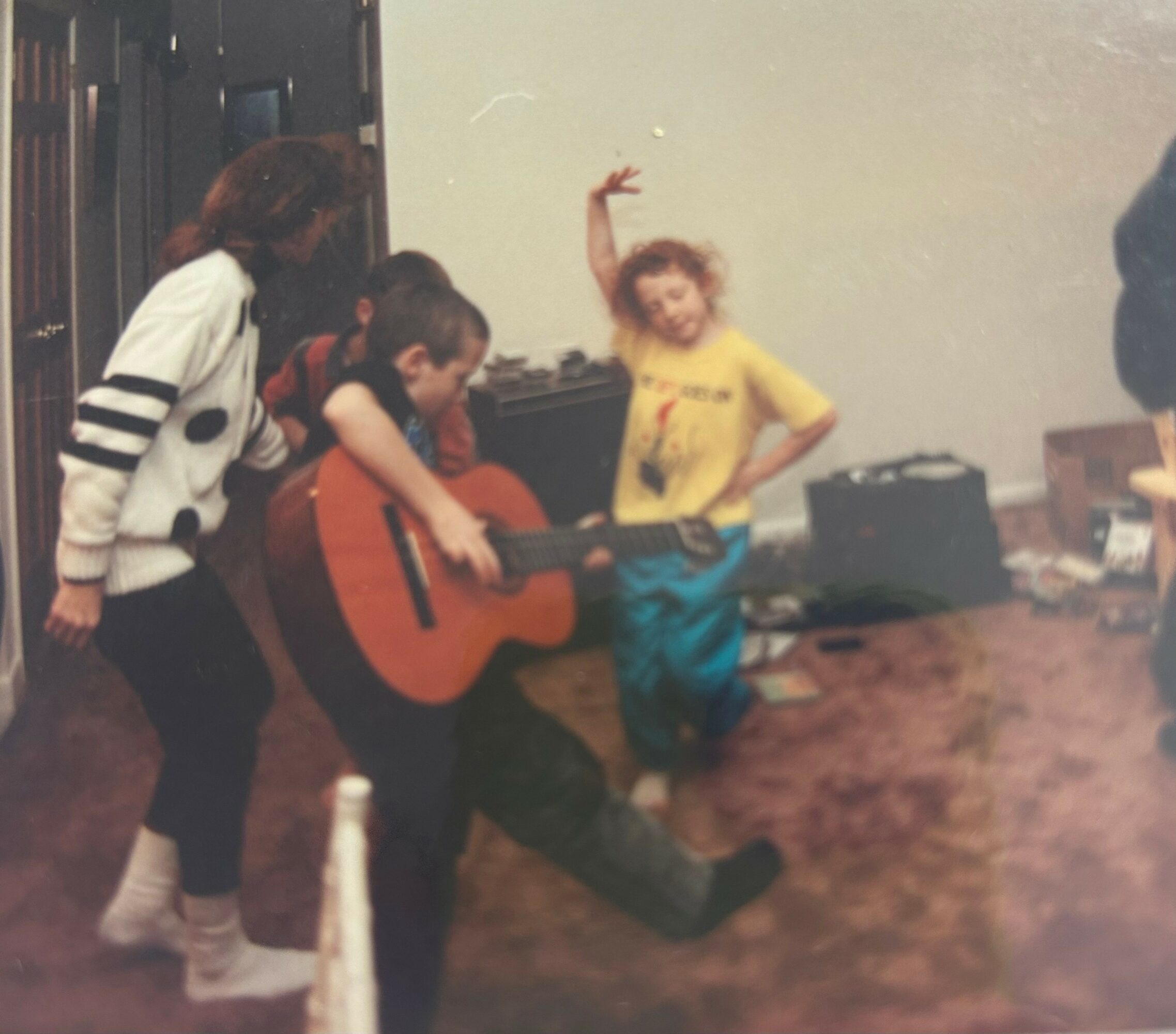Three children and an adult in a room, one child playing guitar, others dancing or watching, with boxes in the background.