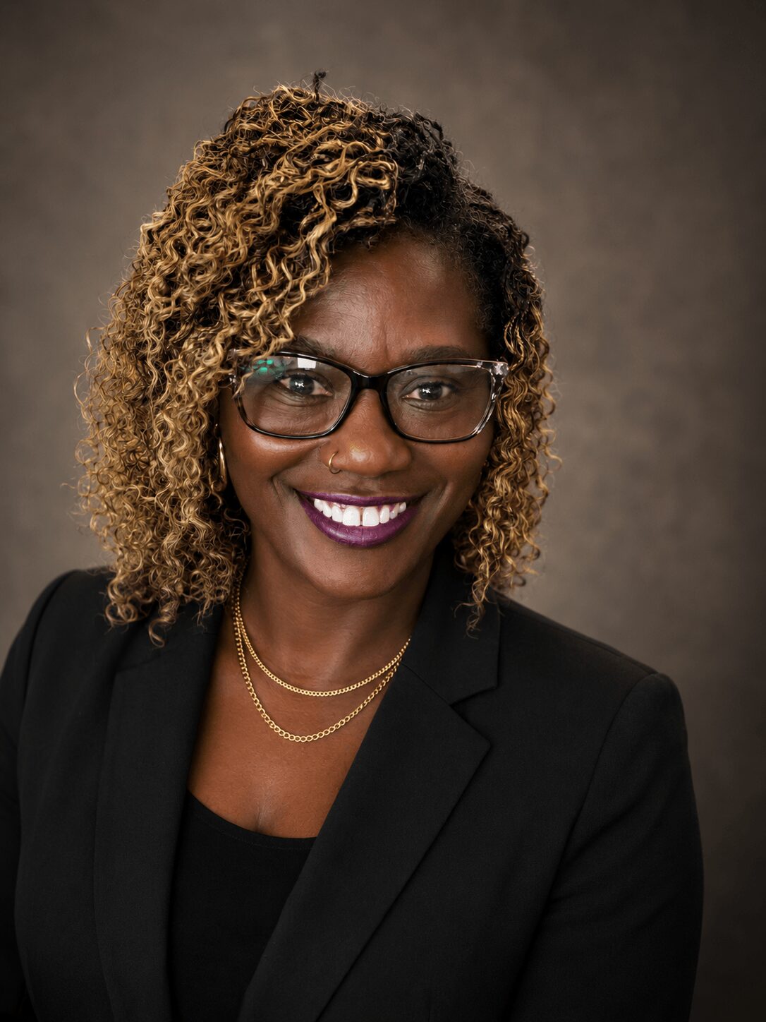 Portrait of a woman with curly hair, glasses, and a black blazer smiling against a neutral background.