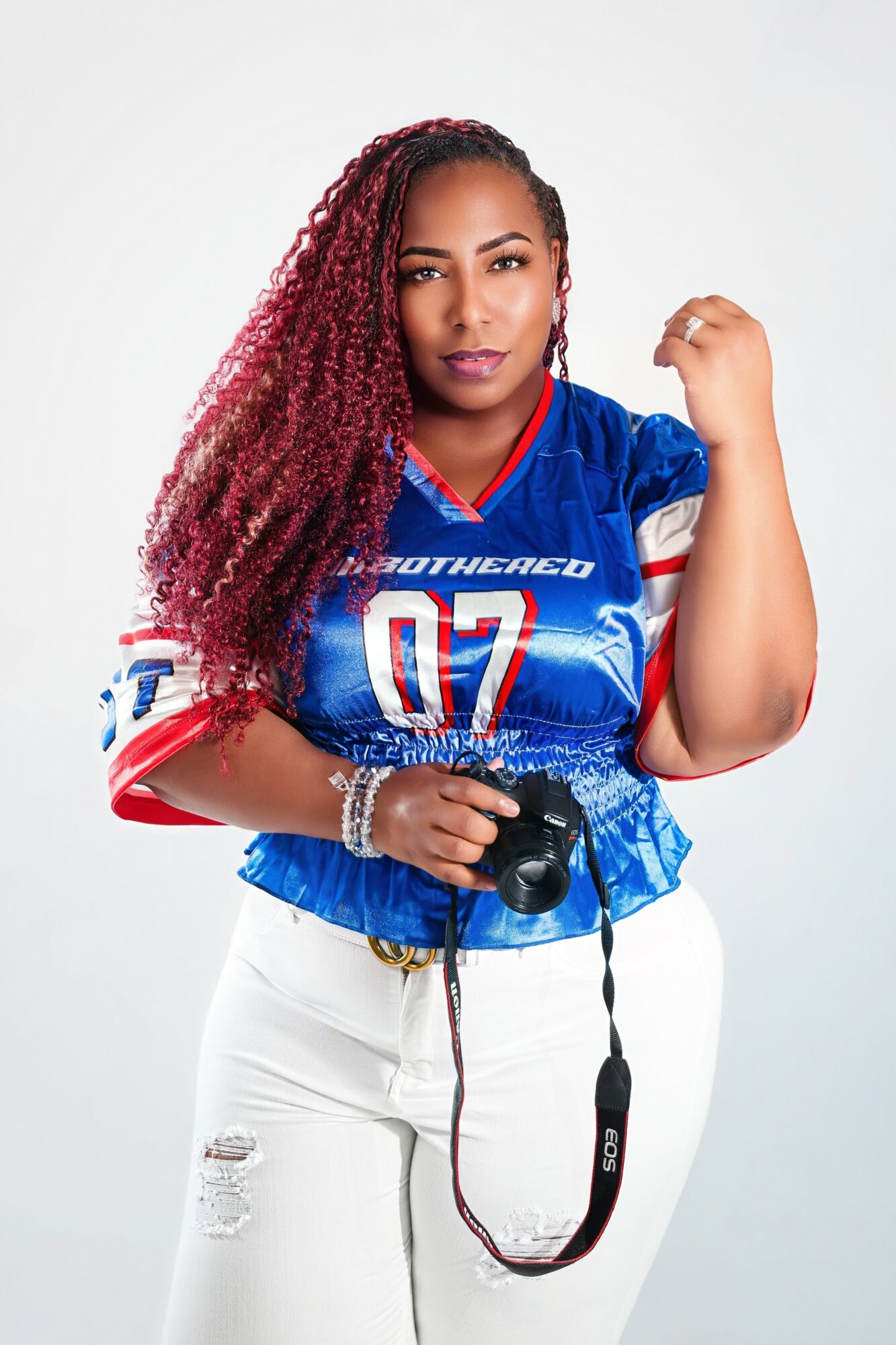 Woman with long, curly red hair wearing a blue sports jersey and white pants, holding a camera, standing against a white background.