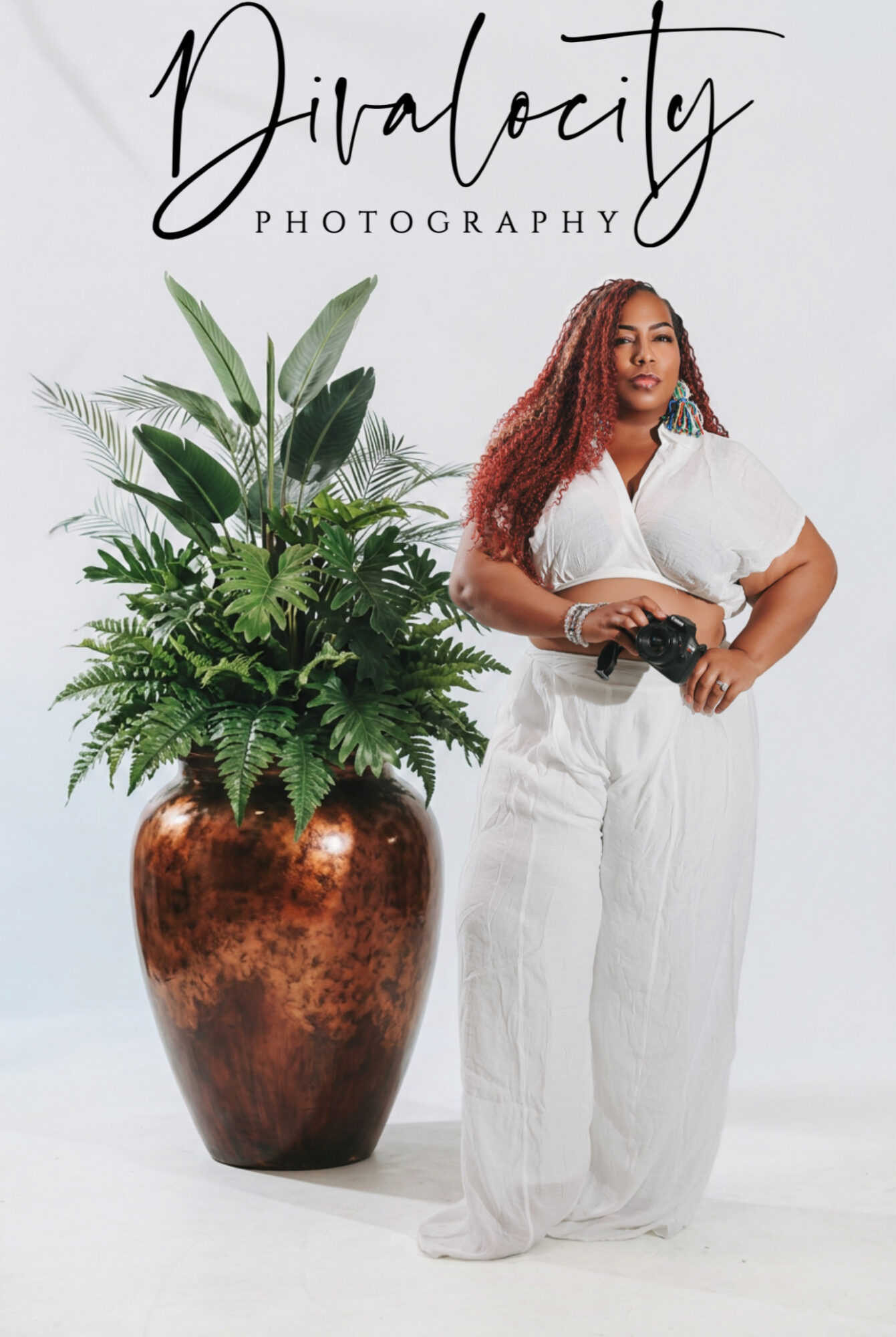 Woman with curly hair holding a camera, standing next to a large potted plant, against a white background.
