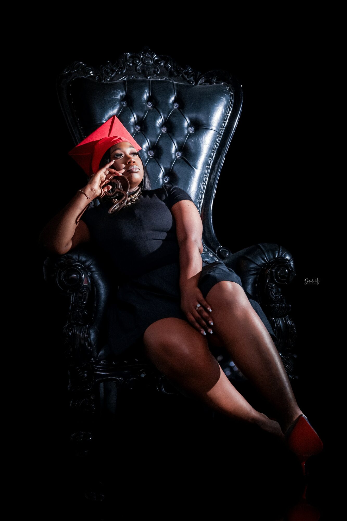 Person sitting on a large, ornate black chair, wearing a red hat and black dress, with one hand near face and legs crossed.