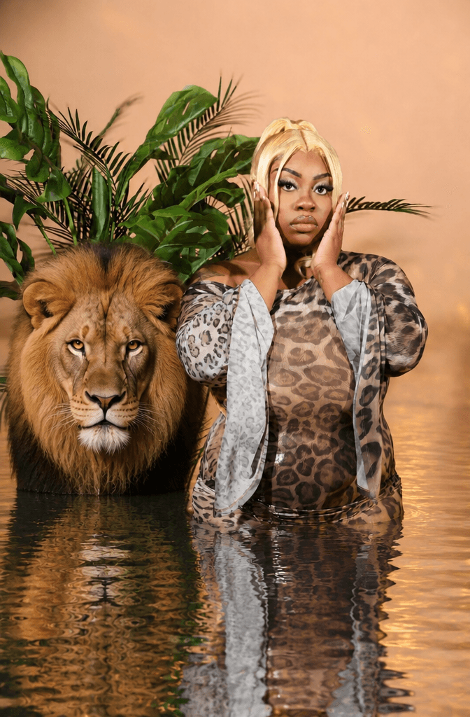 Woman with blonde hair standing in water with a lion and lush green plants behind her, reflecting in water.