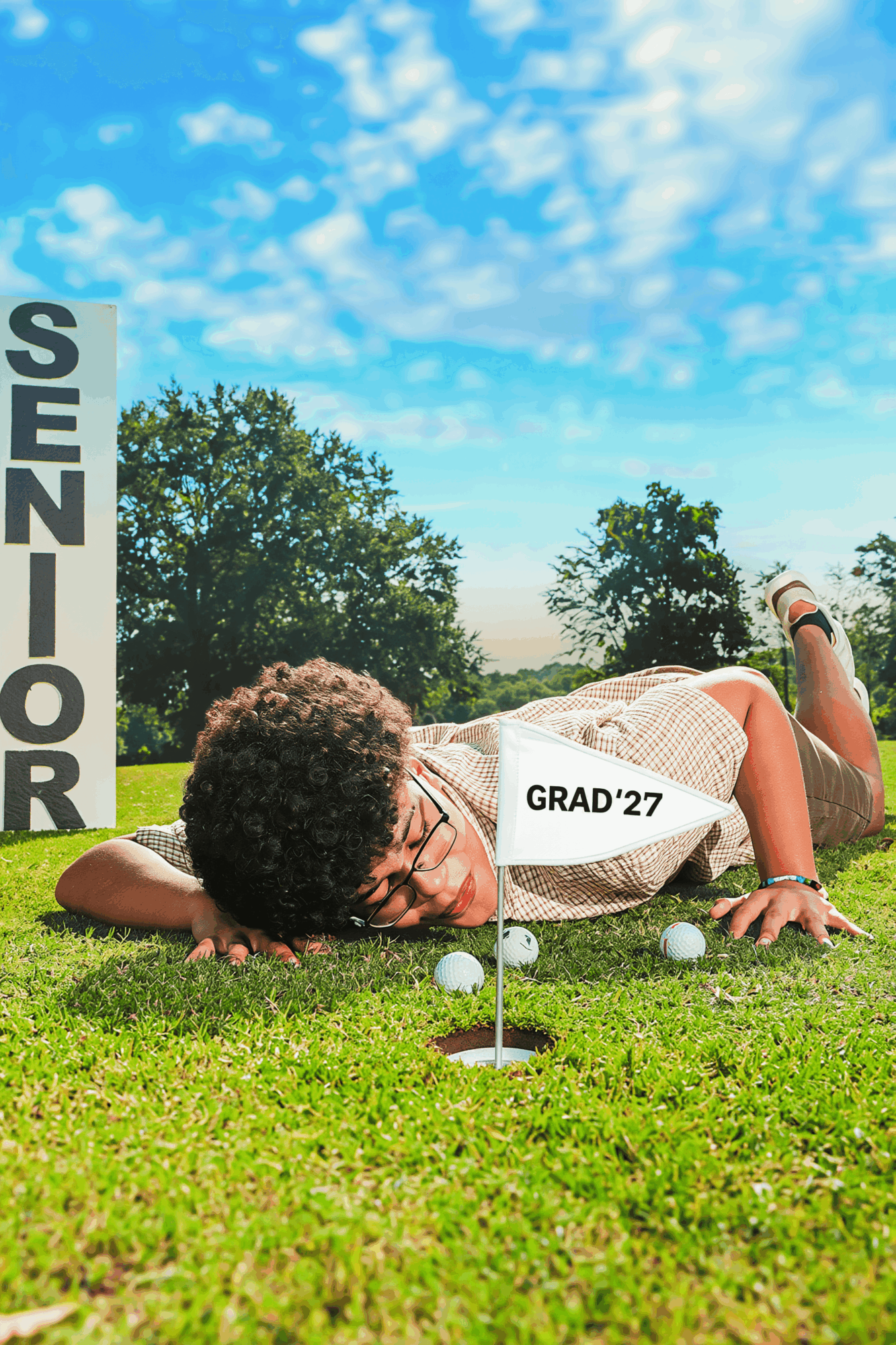Person lying on grass near a golf hole with a sign reading 'SENIOR' and a 'GRAD '27' sash, under a partly cloudy sky.