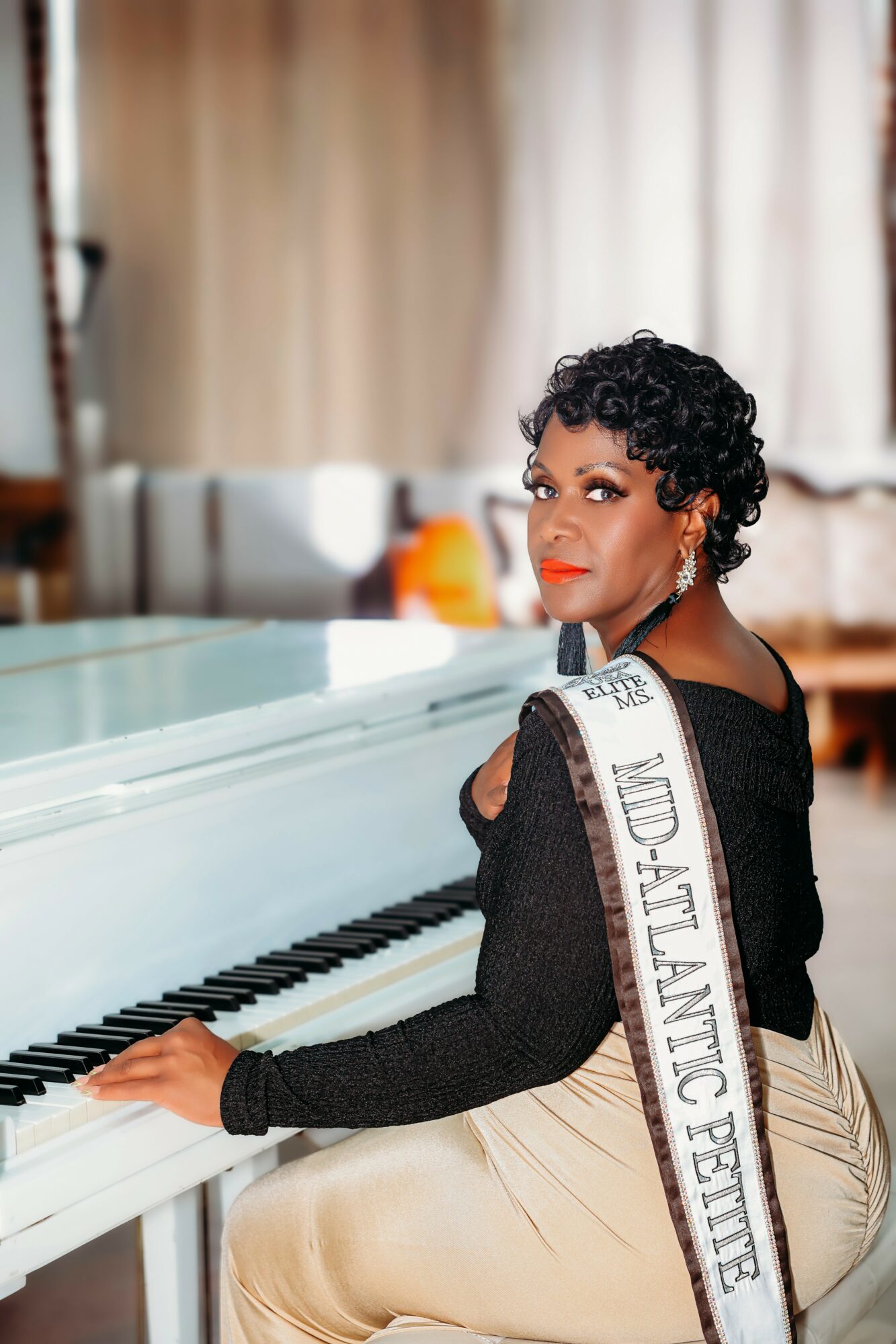 Woman with short curly hair sitting at a white piano, wearing a black top and beige pants, in a room with curtains.