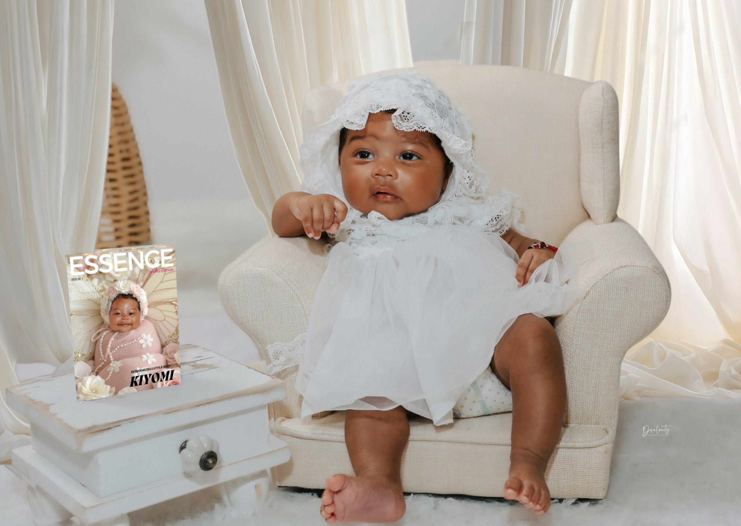 Baby girl in white dress and bonnet sitting on a white armchair, next to a magazine on a small table, in a bright room.
