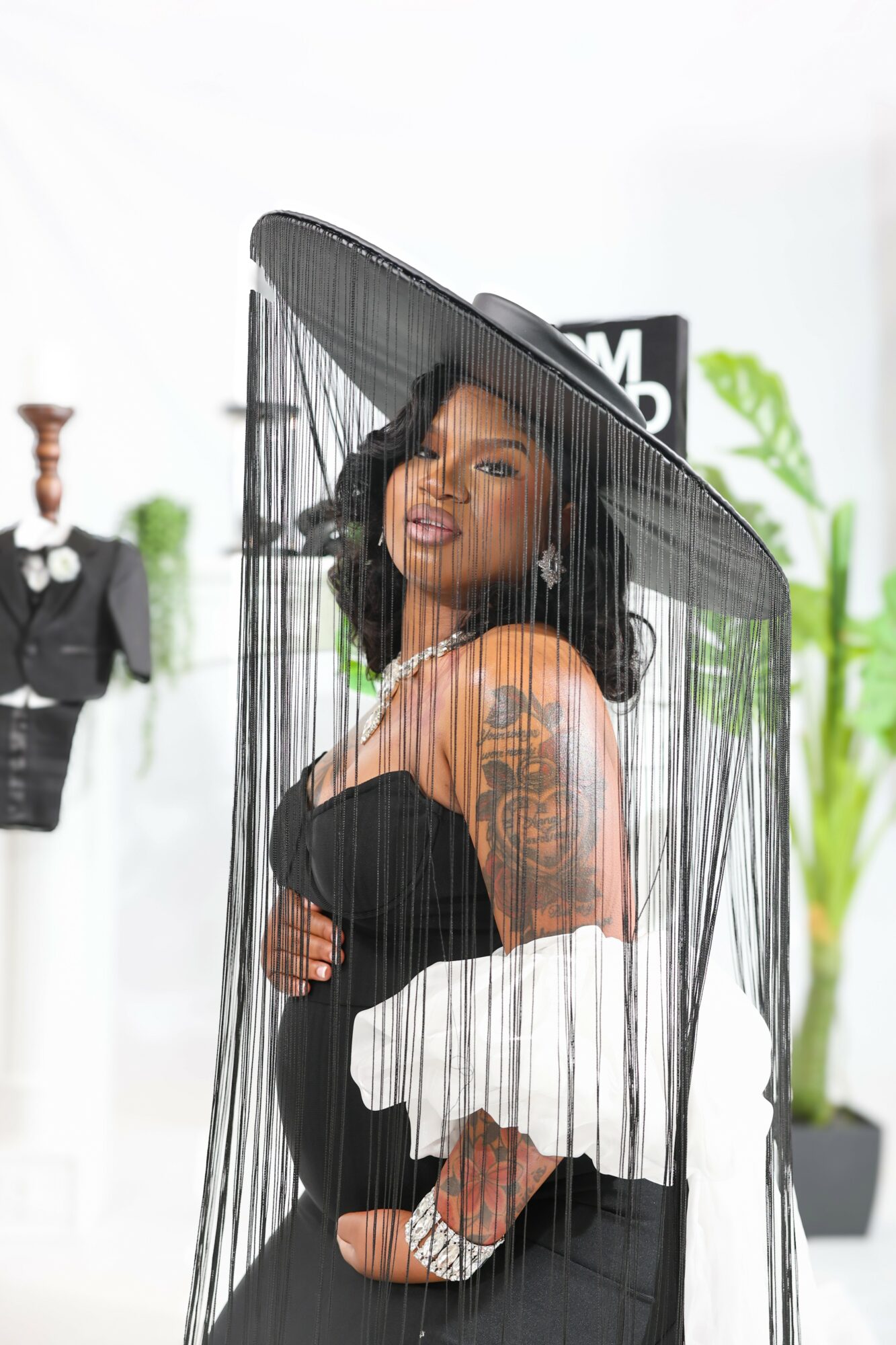 Woman wearing a large black hat with fringe, black dress, and white jacket, standing inside a transparent umbrella, with plants and mannequins in background.