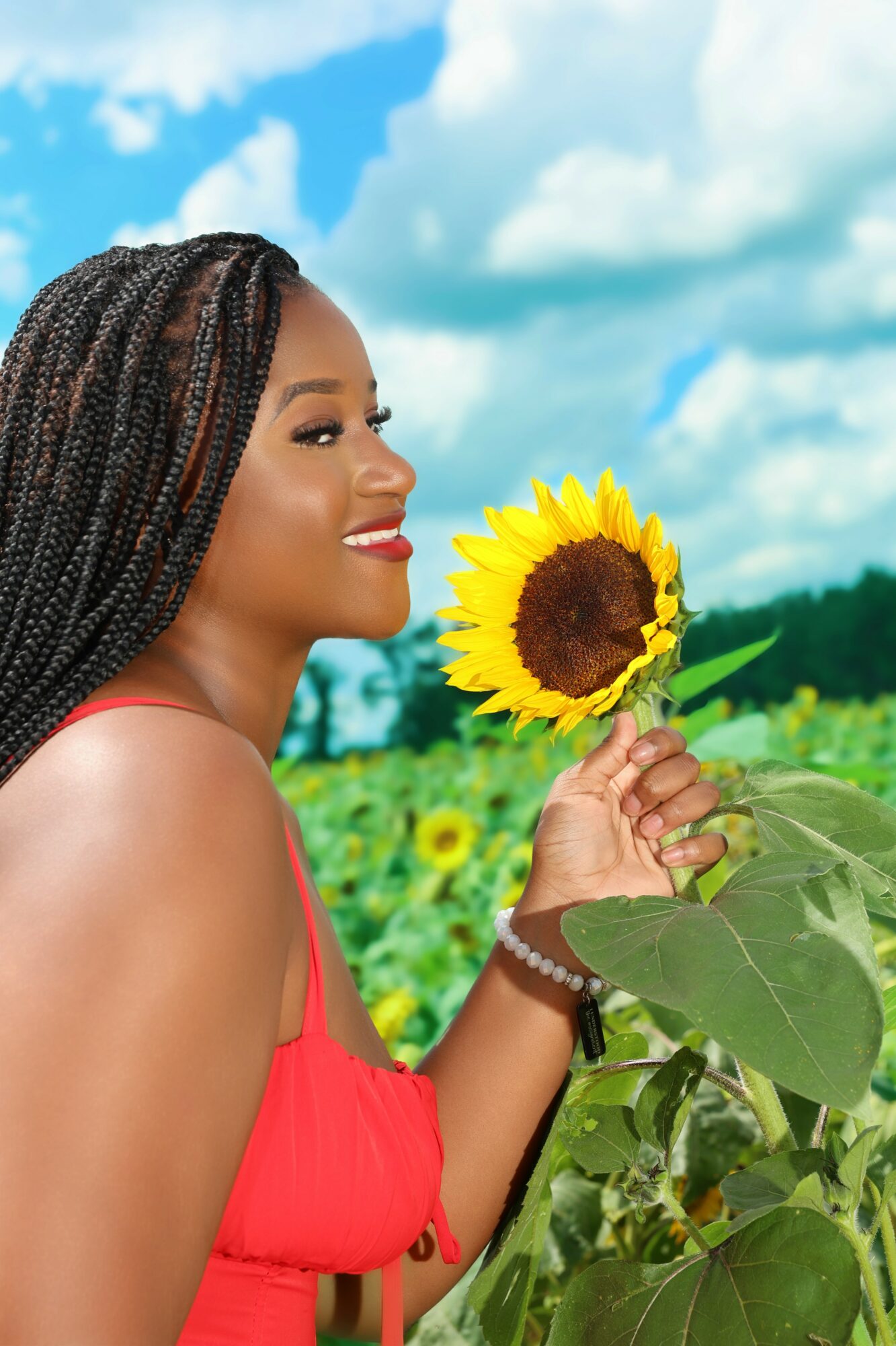 Woman with braided hair holding a sunflower in a field under a blue sky with clouds.